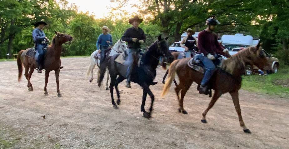 Five riders on horseback in a gravel clearing bordered by trees.